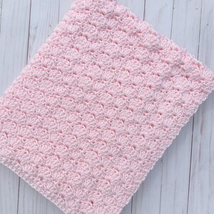 Light pink baby blanket crocheted in a shell stitch which gives it a wonderful texture.  Edges are straight with no border.  Picture taken on a white wooden background.  Blanket is folded in fourths and laid at a forty five degree angle.