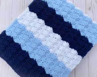May include: A crocheted blanket featuring a repeating ripple pattern with diagonal stripes in navy blue, light blue, and white. The textile has a textured surface created by the shell stitch technique, displaying a consistent color block design.
