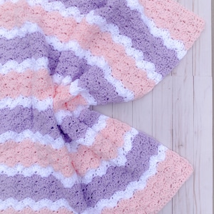 May include: A handmade crochet blanket in a chevron pattern. The blanket features alternating stripes of white, light pink, and lavender. The soft, textured blanket is folded on a light wood surface.