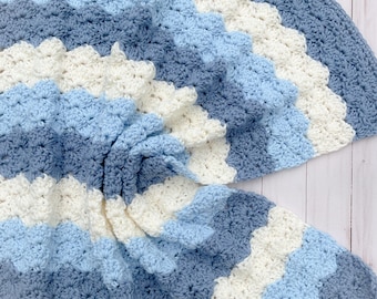 May include: A handmade crochet blanket featuring a repeating shell stitch pattern in alternating horizontal stripes of cream, light blue, and slate blue. The textile has a soft, textured appearance with a scalloped edge design throughout the fabric.