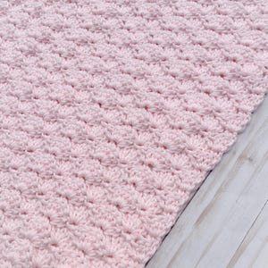 Light pink baby blanket crocheted in a shell stitch which gives it a wonderful texture.  Edges are straight with no border.  Picture taken on a white wooden background.  Blanket is laid out flat and photo taken to show side edge.