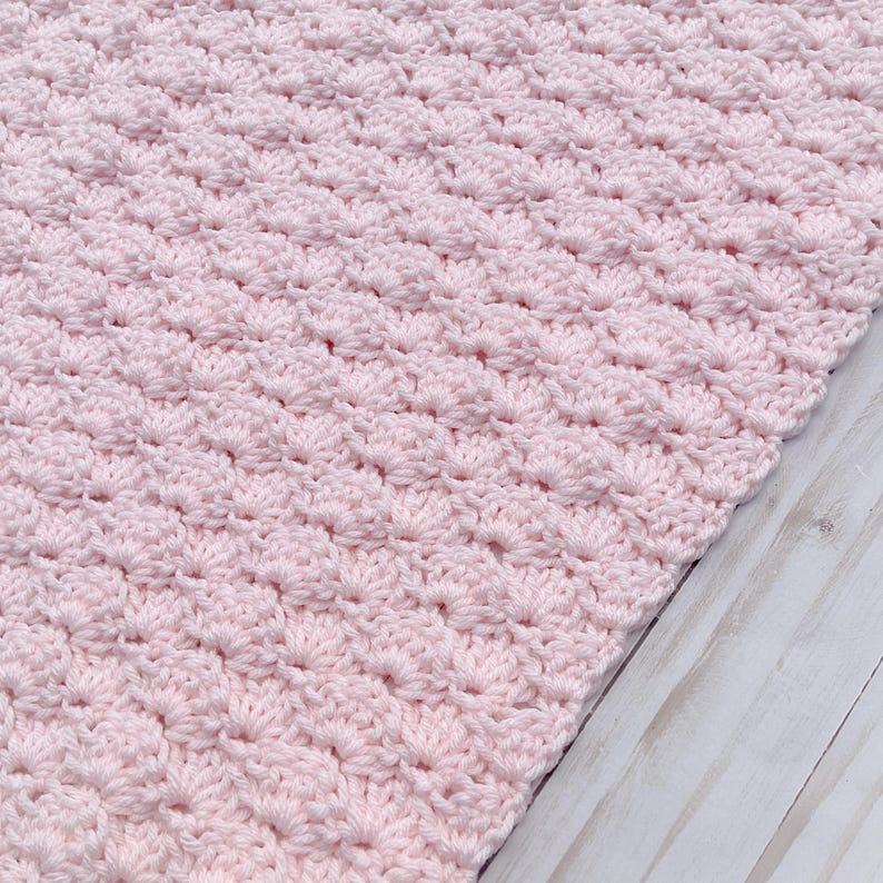 Light pink baby blanket crocheted in a shell stitch which gives it a wonderful texture.  Edges are straight with no border.  Picture taken on a white wooden background.  Blanket is laid out flat and photo taken to show side edge.