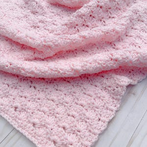 Light pink baby blanket crocheted in a shell stitch which gives it a wonderful texture.  Edges are straight with no border.  Picture taken on a white wooden background.  Picture shows corner of the blanket.