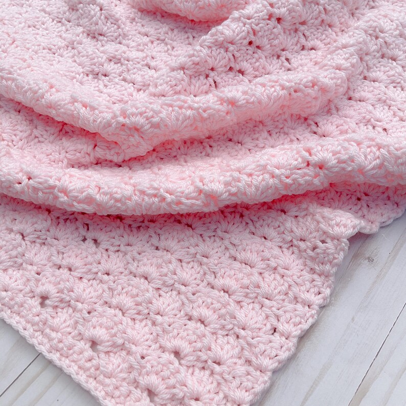 Light pink baby blanket crocheted in a shell stitch which gives it a wonderful texture.  Edges are straight with no border.  Picture taken on a white wooden background.  Picture shows corner of the blanket.