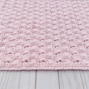 Light pink baby blanket crocheted in a shell stitch which gives it a wonderful texture.  Edges are straight with no border.  Picture taken on a white wooden background.  Blanket is laid out flat and photo taken to show bottom edge.