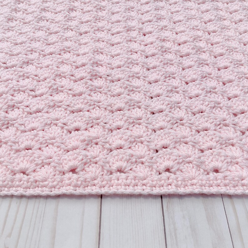Light pink baby blanket crocheted in a shell stitch which gives it a wonderful texture.  Edges are straight with no border.  Picture taken on a white wooden background.  Blanket is laid out flat and photo taken to show bottom edge.