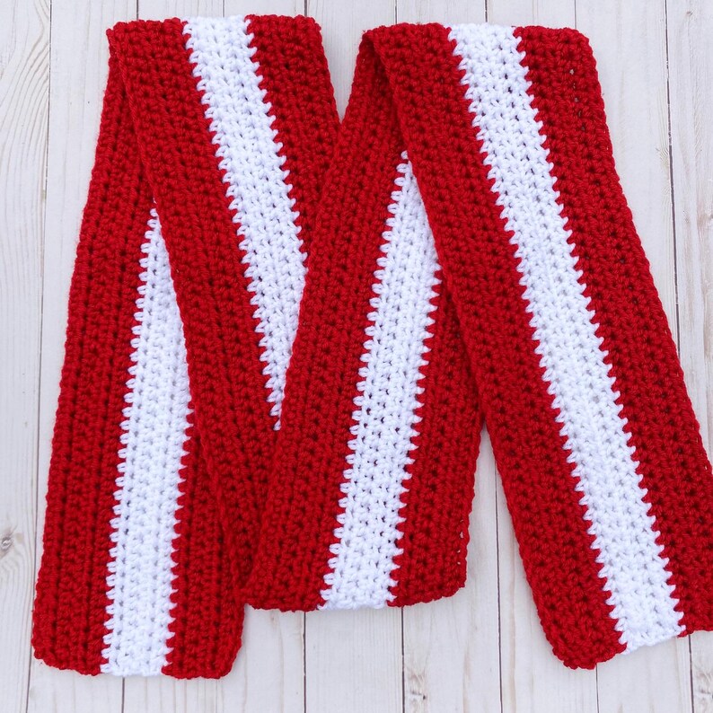 Red and white striped crocheted scarf layed flat and shaped in zig zags.  Stripes run lengthwise on the scarf.  There are two red stripes on the outside lengths of the scarf, and the center stripe is white.