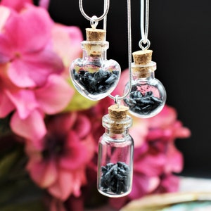 May include: Three glass bottle necklaces with silver chains. Each bottle is filled with black stones and has a cork stopper. The bottles are in the shape of a heart, a circle, and a rectangle.