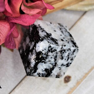 May include: A white and black cube-shaped crystal with a speckled pattern. The crystal is sitting on a light wood surface.