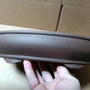 May include: A brown oval ceramic bonsai pot with a slightly raised rim and three small feet.