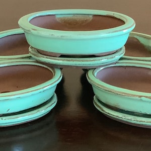 May include: Five oval-shaped, light green ceramic bonsai pots with brown interiors and a light brown rim. The pots are stacked in a pyramid shape.