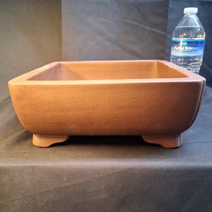 May include: A brown, rectangular bonsai pot with a square shape and four small feet. The pot is made of a textured material and has a wide opening. A bottle of water is in the background.