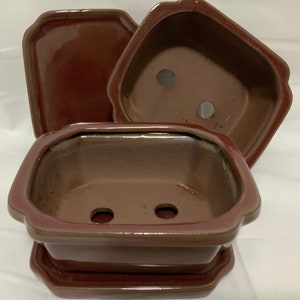 May include: A set of three terracotta-colored ceramic plant pots with drainage holes. The pots are in different sizes and shapes, with a square saucer.