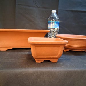 May include: Three terracotta bonsai pots of varying shapes and sizes. The largest is rectangular with flared sides, the middle one is square, and the smallest is oval. A water bottle is in the background.