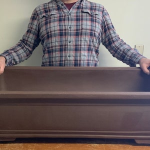 May include: A large, rectangular, brown bonsai pot with a slightly raised rim and a decorative design on the front. The pot is sitting on a wooden surface.