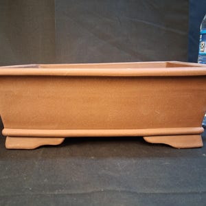 May include: A rectangular, brown bonsai pot with a slightly flared rim and decorative feet. The pot is made of a terracotta-like material and is empty. The pot is approximately 46 cm long.