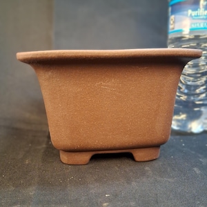 May include: A brown, rectangular bonsai pot with a textured surface. The pot has a slightly flared rim and sits on four small feet. A clear water bottle is in the background.