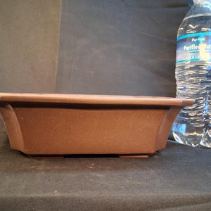 May include: A rectangular brown bonsai pot with a slightly flared design. The pot is made of a matte material and has a low profile. A clear plastic water bottle is in the background.