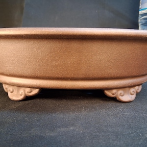 May include: A brown, oval-shaped bonsai pot with a textured surface. The pot has a wide rim and is supported by three decorative feet. The pot is designed for growing bonsai trees.