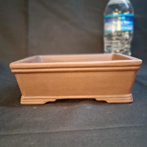 May include: A brown, rectangular bonsai pot with a raised rim and small feet. The pot is empty and sits on a dark surface. A clear plastic water bottle is in the background.