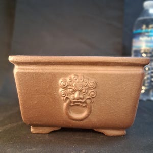 May include: A rectangular brown bonsai pot with a detailed face design on the front. The pot has a textured surface and small feet. The face design includes a ring-shaped handle. Ideal for bonsai enthusiasts.