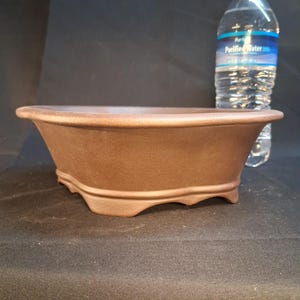 May include: A brown, rectangular bonsai pot with a flared shape and curved feet. The pot is made of ceramic and has a smooth, matte finish. A clear plastic water bottle is in the background.