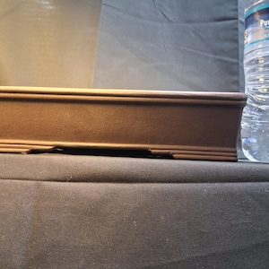 May include: A rectangular brown ceramic bonsai pot with a textured surface. The pot has a raised rim and decorative detailing along the bottom edge. A clear plastic water bottle is next to the pot.