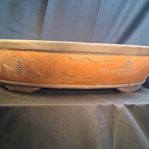 May include: A brown ceramic bonsai pot with a rectangular shape and rounded edges. The pot features a dark rim and is decorated with etched landscape designs, including trees and mountains. It rests on three small feet.