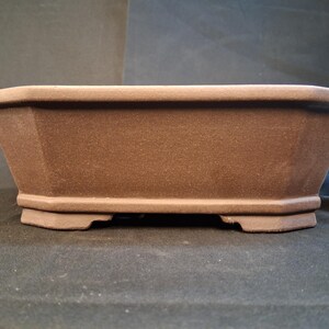 May include: A brown, rectangular bonsai pot with a textured surface. The pot has a wide, octagonal shape with a flat base and is designed for planting small trees or shrubs. A water bottle is in the background.