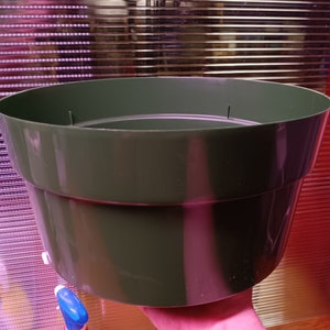 May include: A dark green plastic plant pot with a wide, slightly flared design. The pot is held by a hand, showcasing its size and shape. The background features a textured, reflective surface.