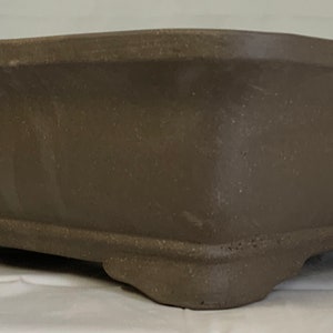 May include: A brown rectangular ceramic bonsai pot with four feet. The pot has a textured surface and a slightly rounded edge.