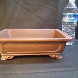 May include: A rectangular brown bonsai pot with decorative feet and a slightly raised rim. The pot is empty and sits on a dark surface. A clear plastic water bottle is next to the pot.