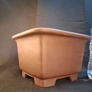 May include: A square, brown bonsai pot with a slightly textured surface. The pot has a wide, curved rim and is supported by four small, square feet. The pot is designed for growing bonsai trees.