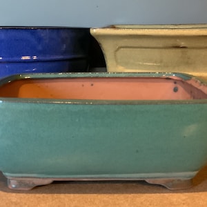 以下が含まれることがあります： 異なる色とサイズのセラミック盆栽鉢が3つあります。最大の鉢は明るい黄色、中くらいの鉢は緑がかった青色、最小の鉢は濃い青色です。