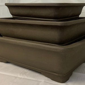 May include: Three brown rectangular bonsai pots of varying sizes are stacked. The pots have a slightly textured surface and a classic, understated design. The largest pot is at the bottom, with the smallest on top, creating a tiered display.