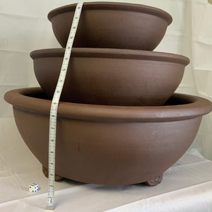 May include: Three brown ceramic pots of varying sizes stacked on top of each other. The pots have a rounded shape and a slightly flared lip. A white measuring tape is visible in the image.