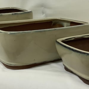 May include: Three rectangular ceramic bonsai pots in various sizes. The pots are a light beige color with a brown rim and a dark glaze on the inside.
