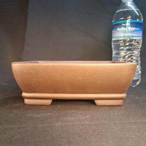 May include: A rectangular, brown bonsai pot with a textured surface. The pot has a low profile and sits on four small feet. A clear plastic water bottle is in the background.