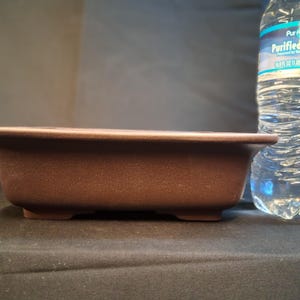 May include: A rectangular brown bonsai pot with a textured surface and a slightly flared rim. The pot sits on four small feet. A clear plastic water bottle is in the background.