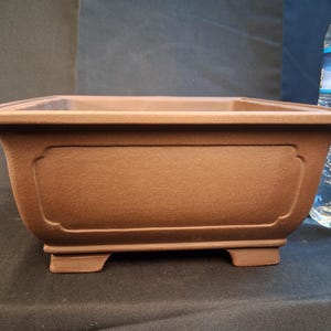 Puede incluir: Una maceta rectangular marrón para bonsái con una superficie texturizada. La maceta tiene un diseño ancho y poco profundo con un borde ligeramente ensanchado y paneles decorativos en los lados. Se apoya sobre cuatro pequeños pies.