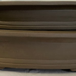 May include: Two brown ceramic bonsai pots, one stacked on top of the other. The pots are oval shaped with a raised rim and a decorative groove around the center.