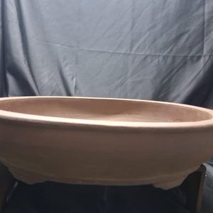 May include: A shallow, oval-shaped terracotta bonsai pot with a smooth, matte finish and a wide, rounded rim. The container features a natural, earthy brown color and is supported by small, integrated feet at the base, providing a stable foundation.