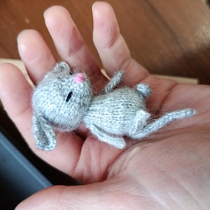 May include: A small, handmade gray knitted bunny with black eyes and a pink nose. The bunny is lying on its side, with its ears and limbs extended. The toy is held in a person's hand.