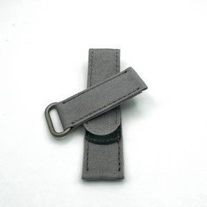 May include: A gray canvas watch strap with a metal buckle. The strap is folded in half and the buckle is visible.