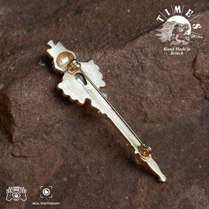 May include: Gold-toned key-shaped brooch with a large, clear crystal in the center. The brooch is detailed with intricate designs and has a pin on the back. The brooch is on a brown, textured surface. The text "TIME GALLERY" and "Hand Made in British" is visible in the background.