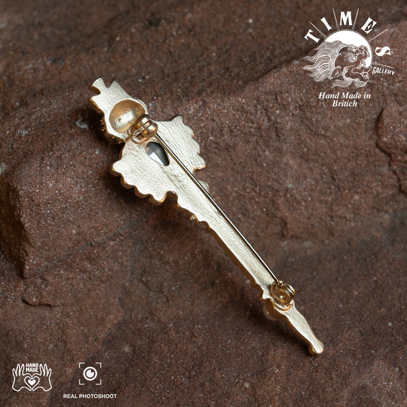 May include: Gold-toned key-shaped brooch with a large, clear crystal in the center. The brooch is detailed with intricate designs and has a pin on the back. The brooch is on a brown, textured surface. The text "TIME GALLERY" and "Hand Made in British" is visible in the background.