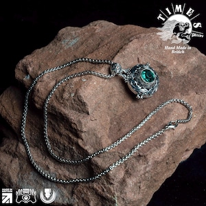 May include: A silver chain necklace with a pendant featuring an ornate design and a large, green gemstone. The necklace is displayed on a rough, brown rock. The text "TIMES GALLERY" and "Hand Made in British" is visible in the background.