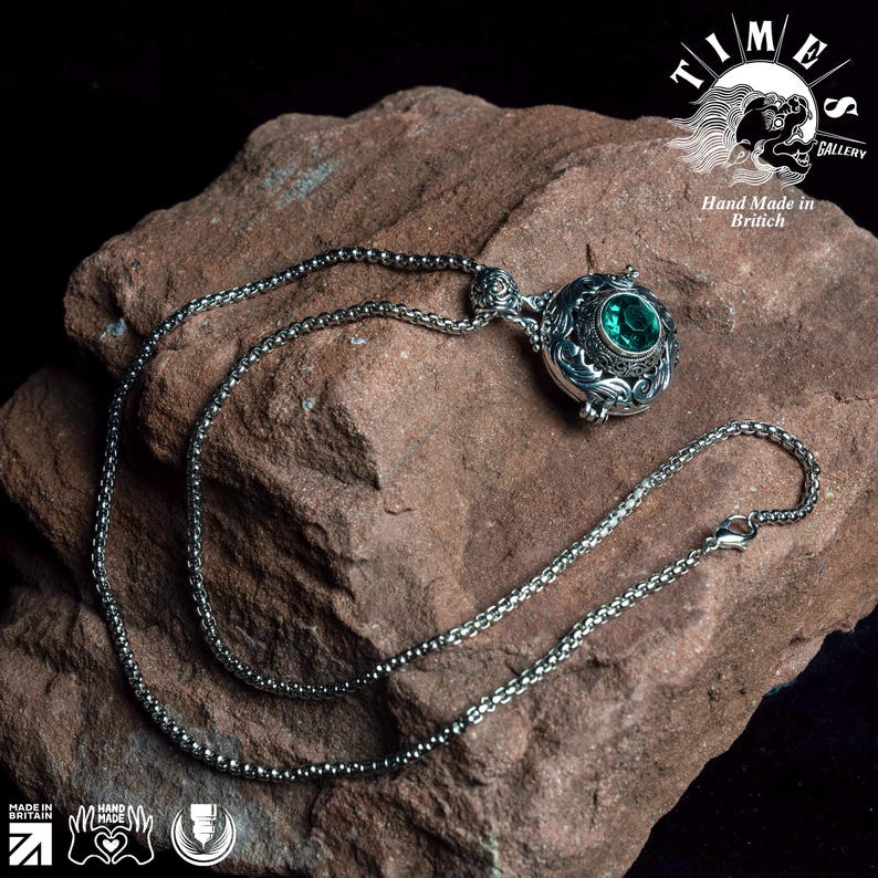 May include: A silver chain necklace with a pendant featuring an ornate design and a large, green gemstone. The necklace is displayed on a rough, brown rock. The text "TIMES GALLERY" and "Hand Made in British" is visible in the background.