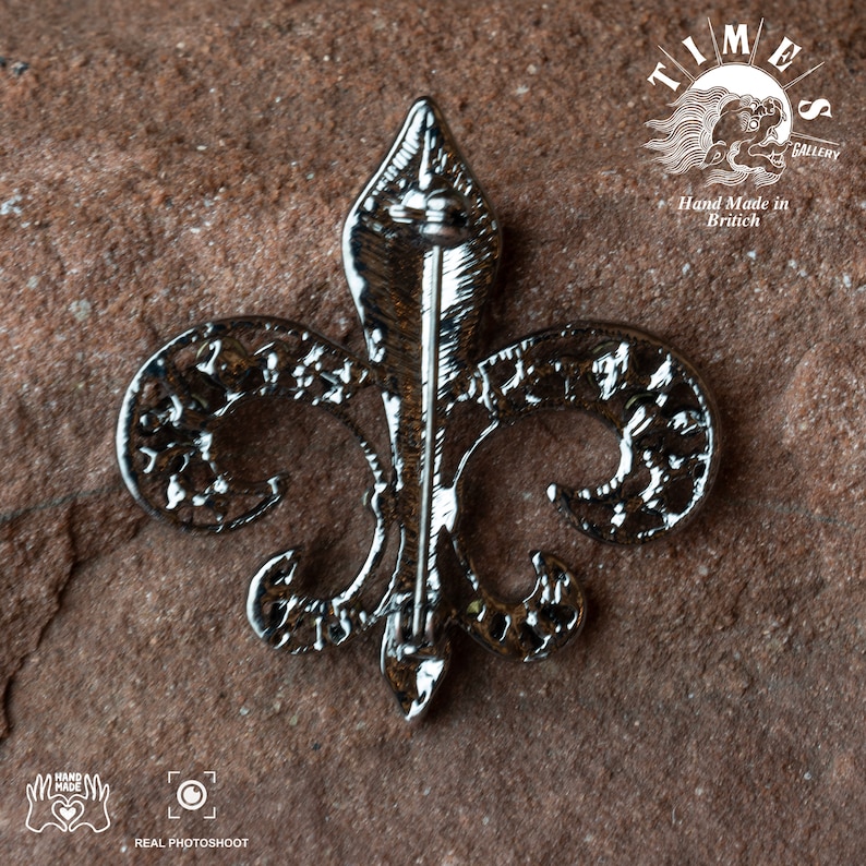 May include: Silver-toned fleur-de-lis brooch with intricate details. The brooch has a pin back and is made in Britain.