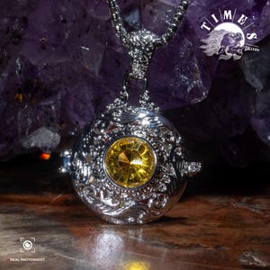 May include: A silver pendant necklace with a round, ornate cage design. The cage contains a yellow gemstone. The necklace is on a purple background.
